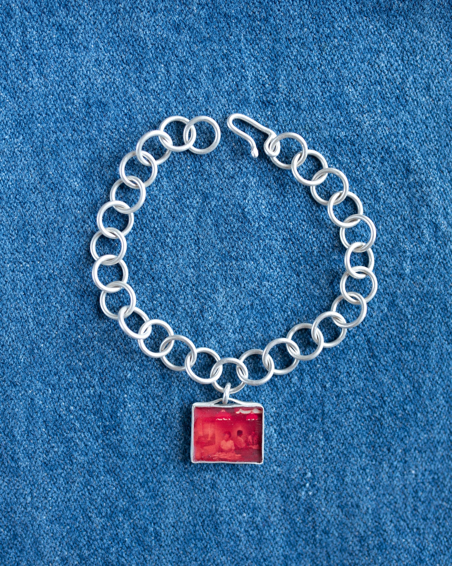 A silver chain bracelet with a square red pendant, displayed on a textured blue fabric background.