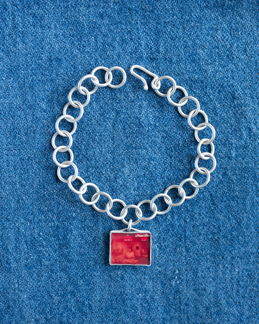 A silver chain bracelet with a square red pendant, displayed on a textured blue fabric background.