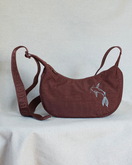A mahogany crescent-shaped shoulder bag with a matching adjustable strap, featuring a white embroidered koi fish design on the front and 'BAGGU' embossed on the strap, set against a light gray fabric backdrop.