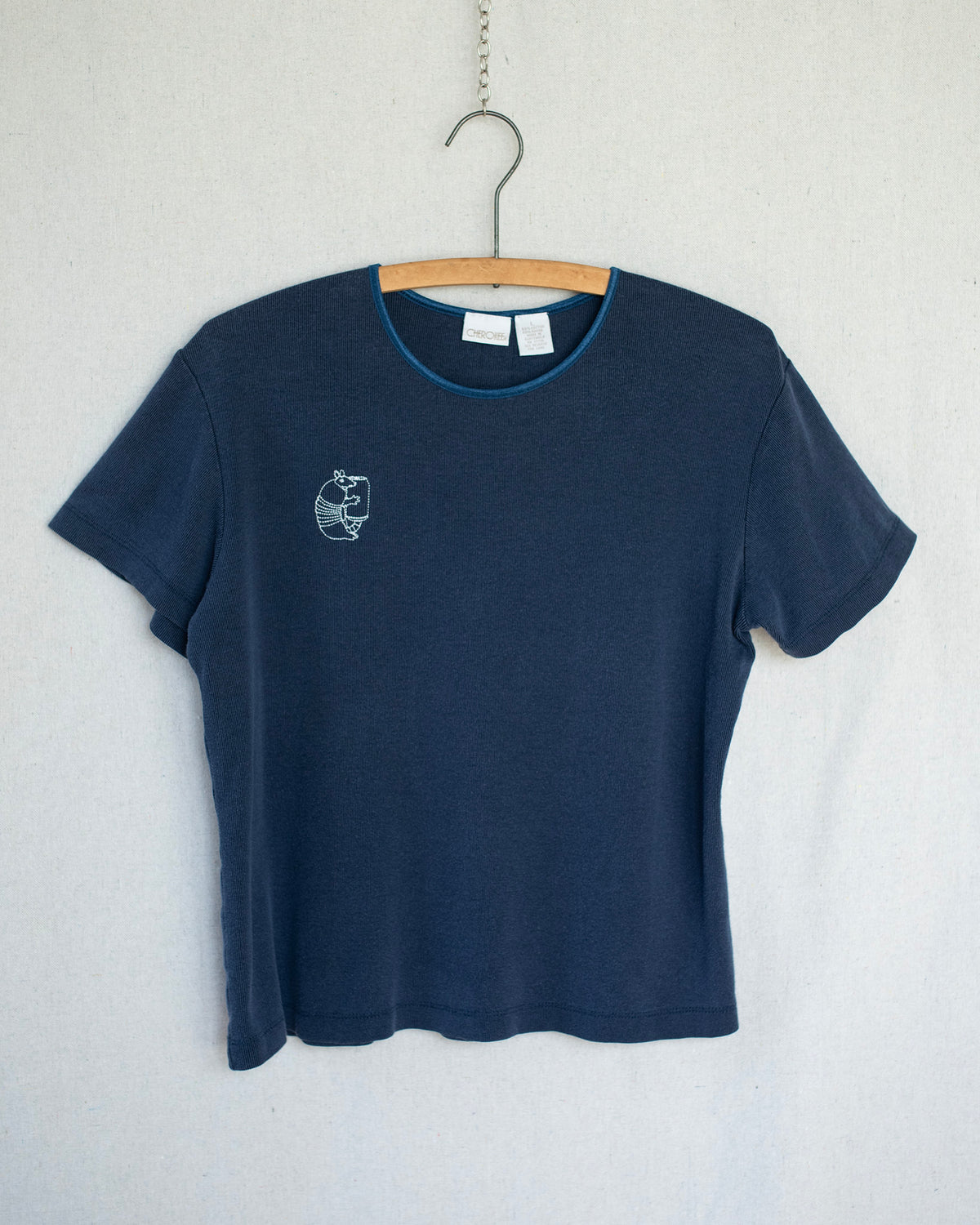 A dark navy blue short-sleeve t-shirt hanging on a wooden hanger against a light gray textured wall. The shirt features a small white embroidery of an armadillo on the left chest and a thin light blue inner collar trim.