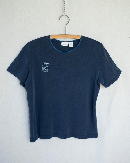 A dark navy blue short-sleeve t-shirt hanging on a wooden hanger against a light gray textured wall. The shirt features a small white embroidery of an armadillo on the left chest and a thin light blue inner collar trim.