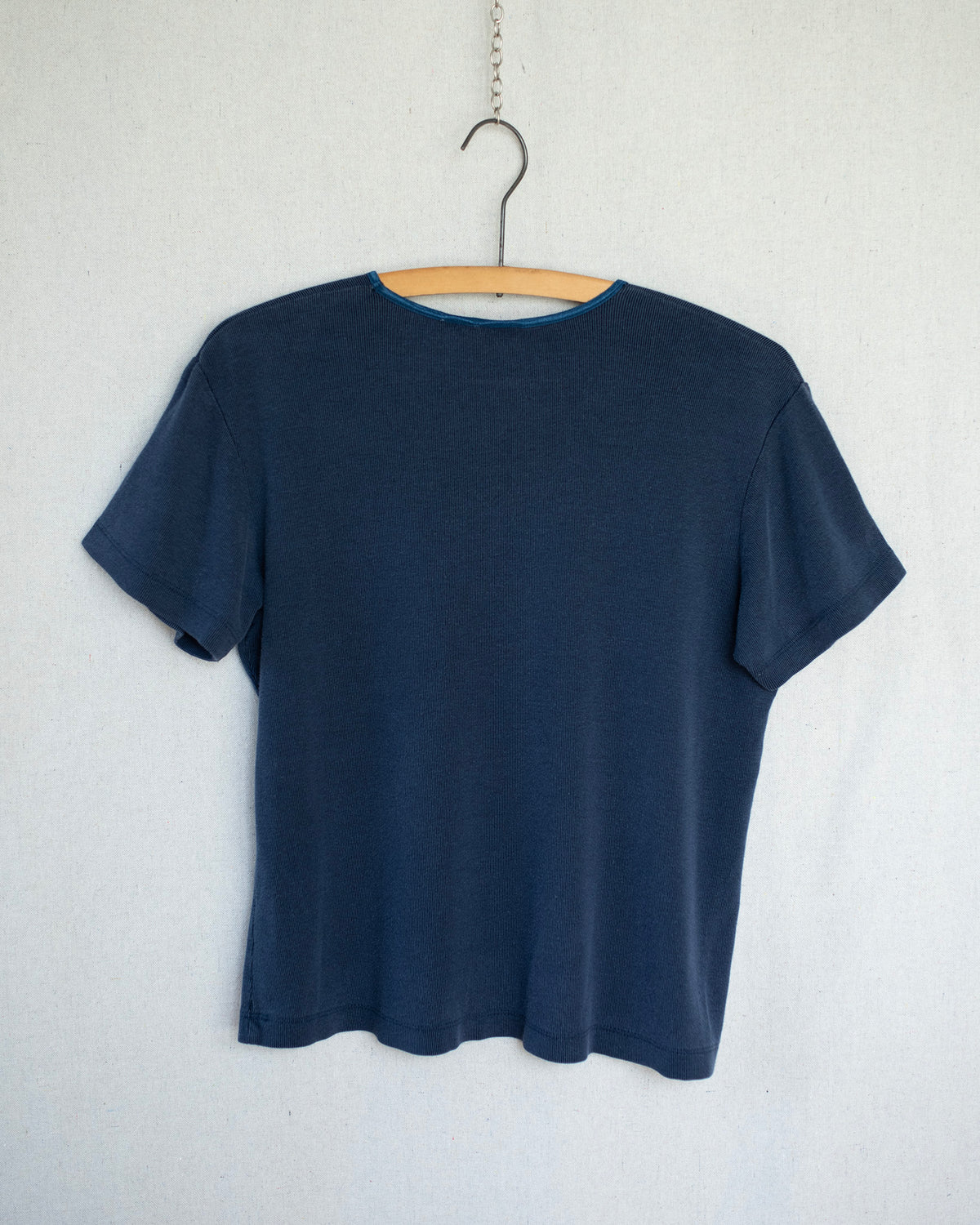 A dark navy blue short-sleeve t-shirt hanging on a wooden hanger against a light gray textured wall.