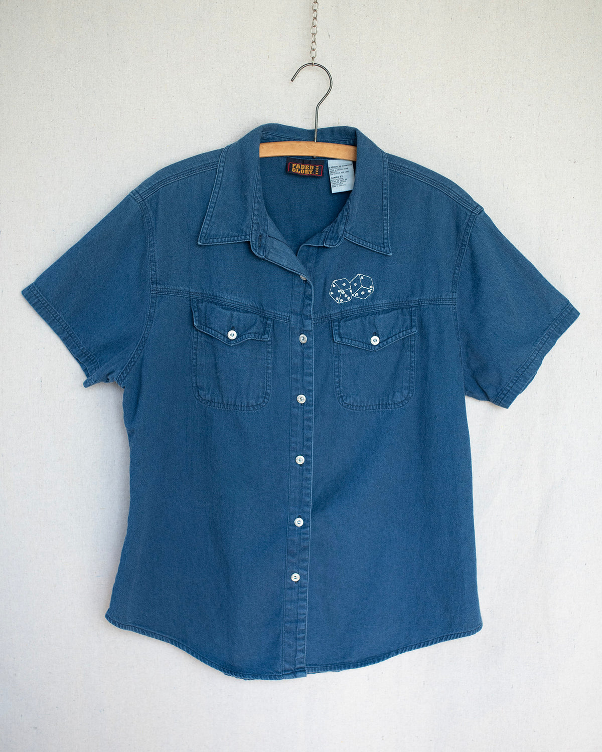 Denim short sleeve shirt hanging on a wooden hanger and has a small dice embroidery above the chest pocket.