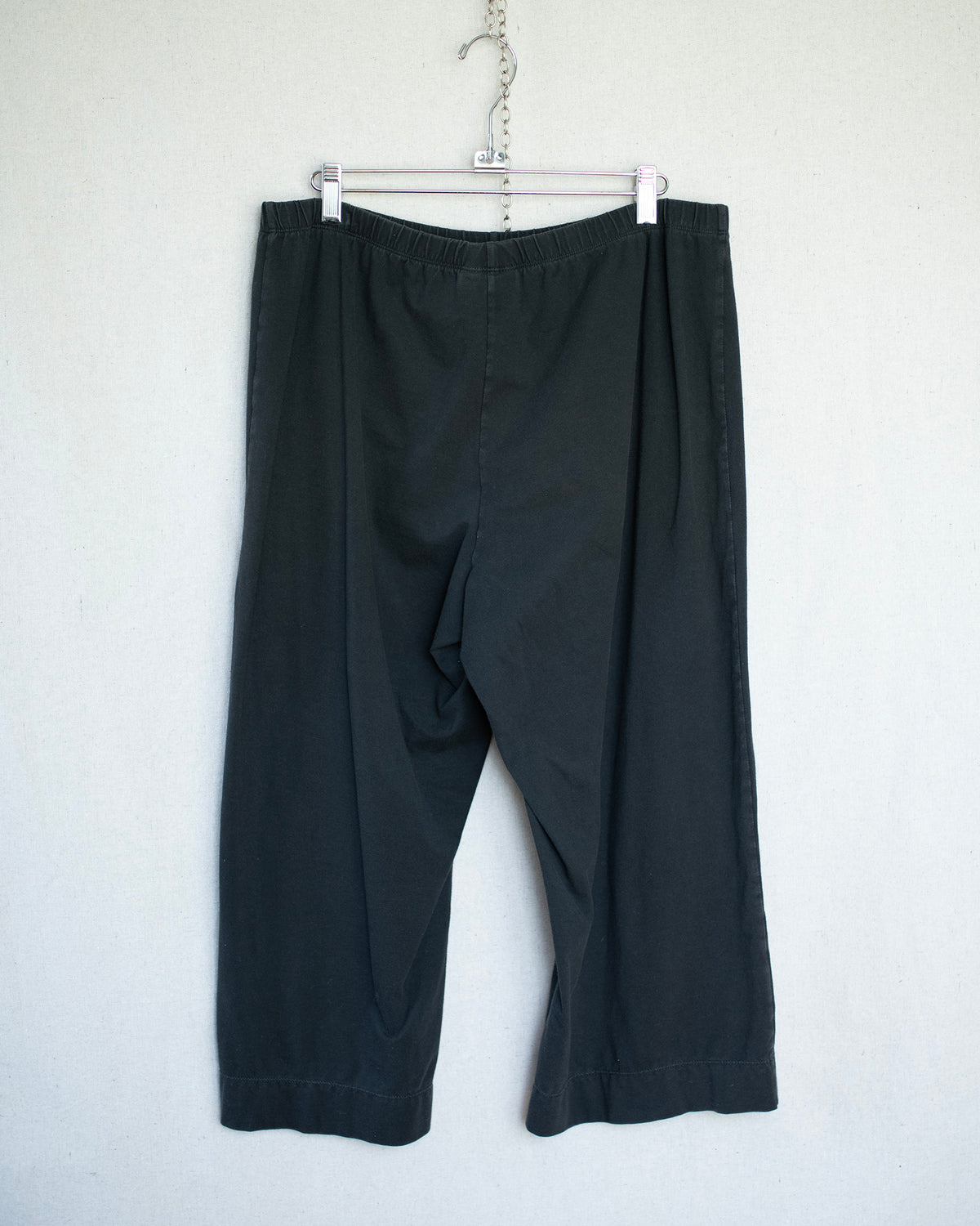 Black cropped pants hanging from a metal hanger against a plain white wall.