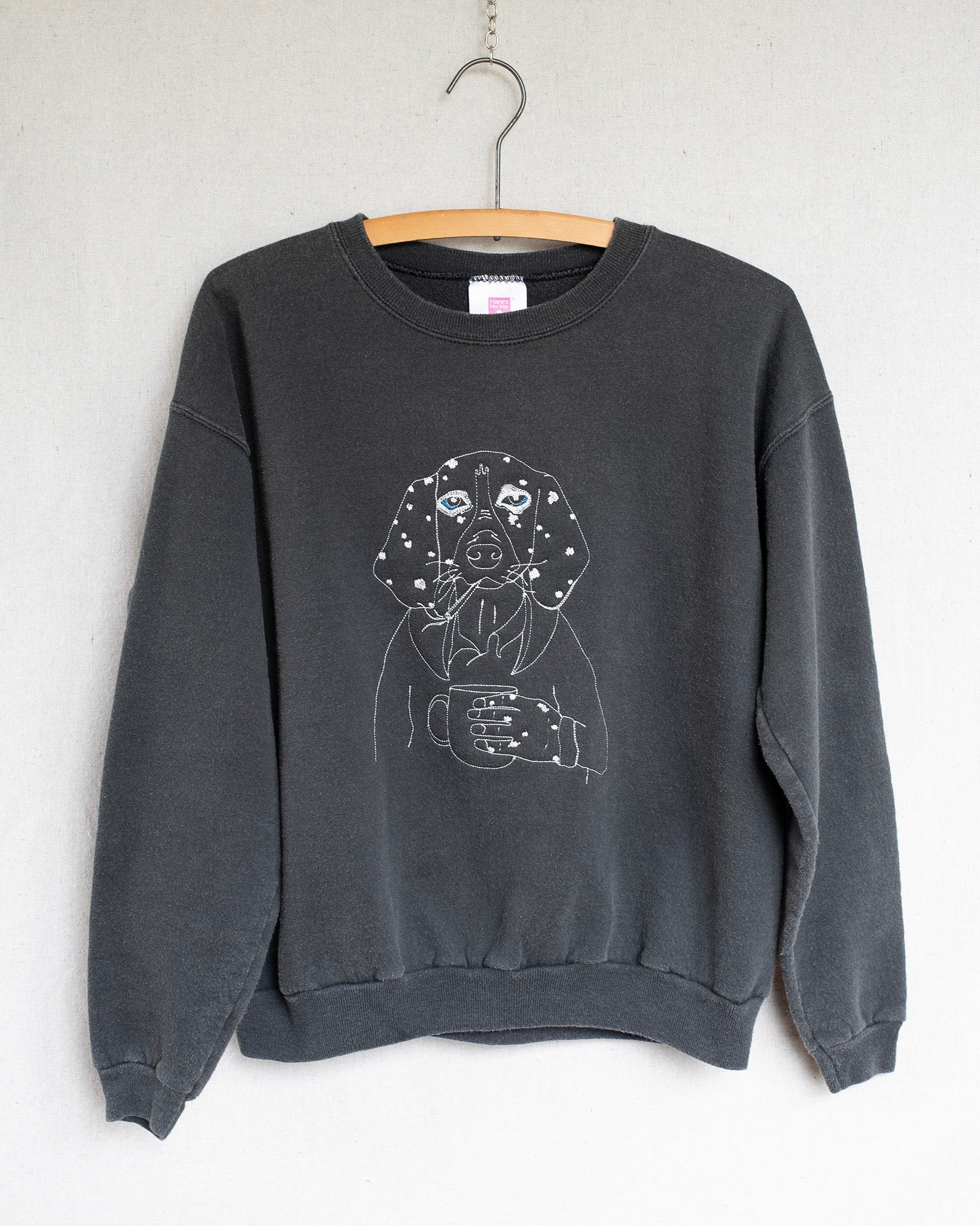 Black sweatshirt with an embroidery of a dog holding a coffee cup on a hanger against a white wall.