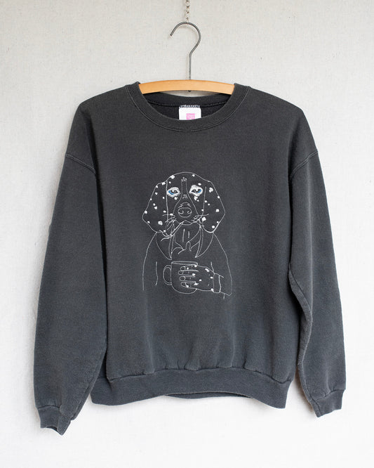 Black sweatshirt with an embroidery of a dog holding a coffee cup on a hanger against a white wall.