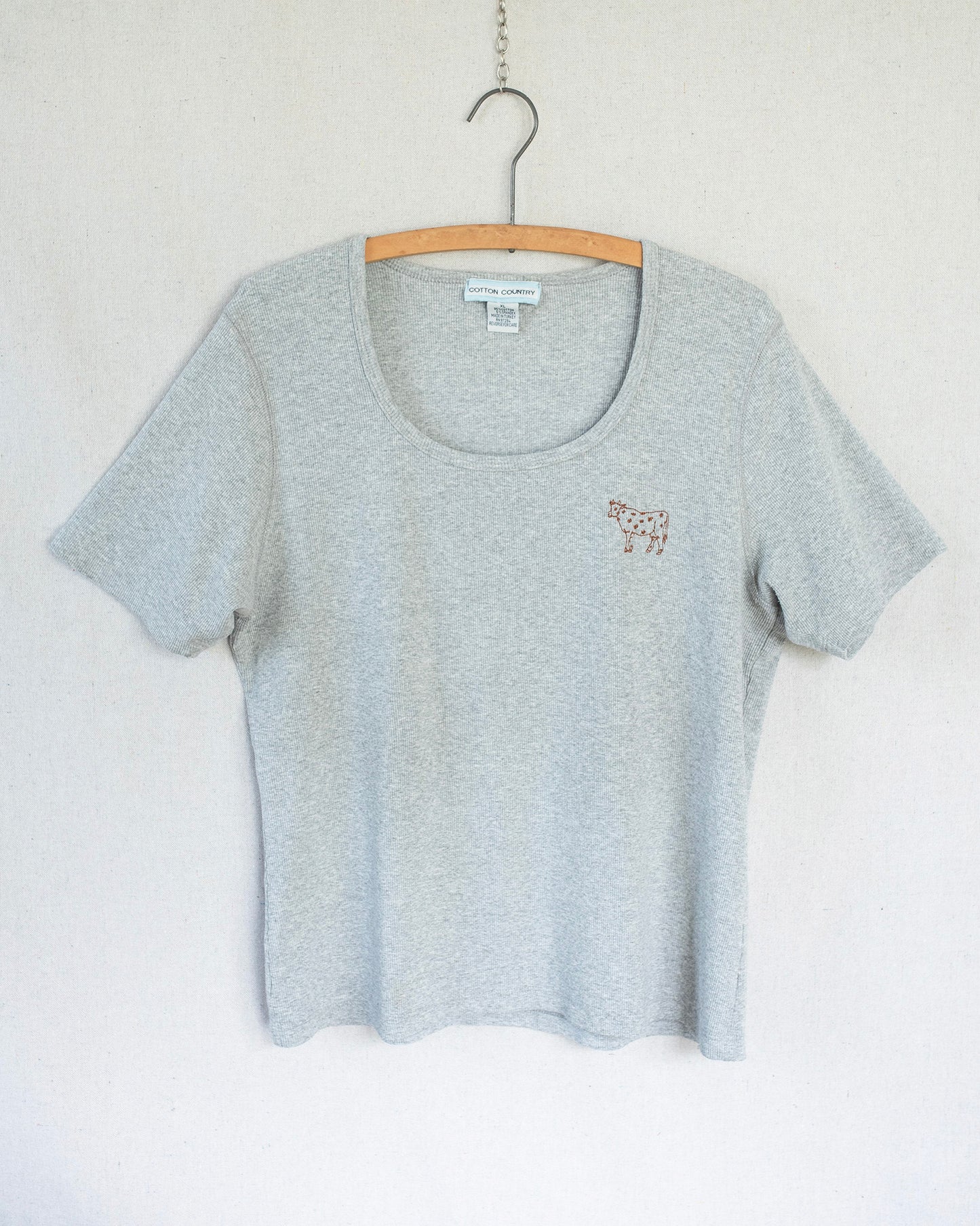Grey t-shirt with a small cow embroidery hanging against a white background