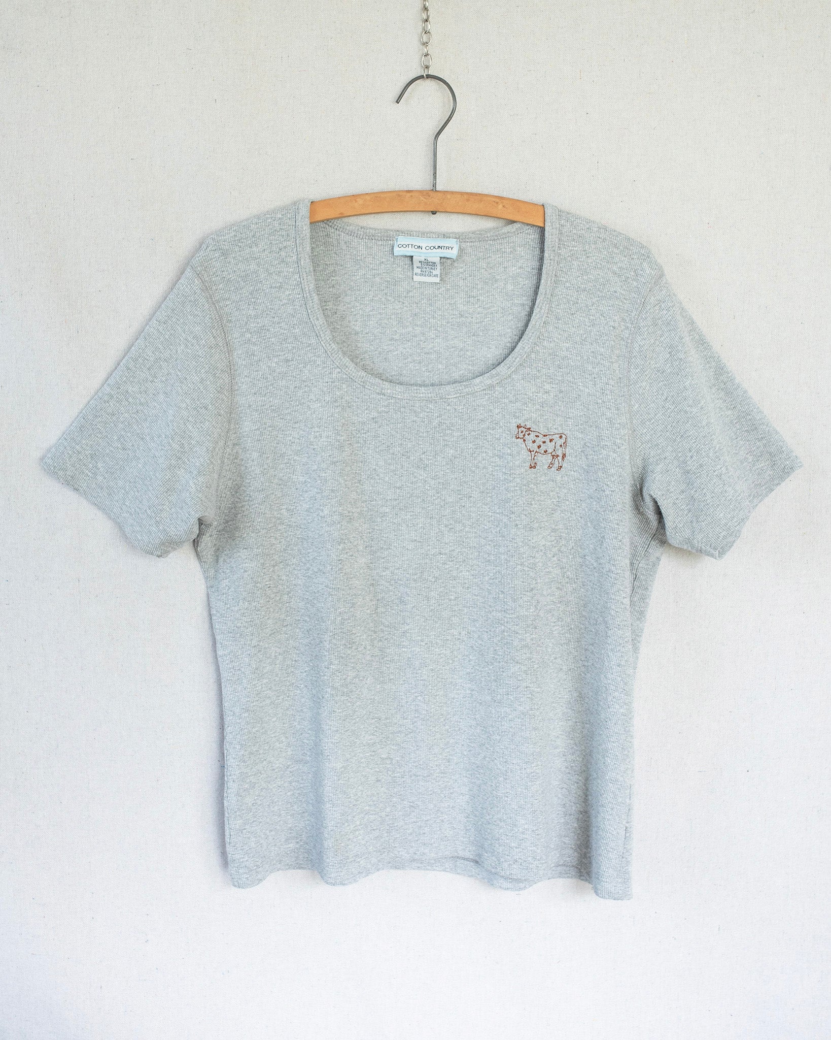 Grey t-shirt with a small cow embroidery hanging against a white background