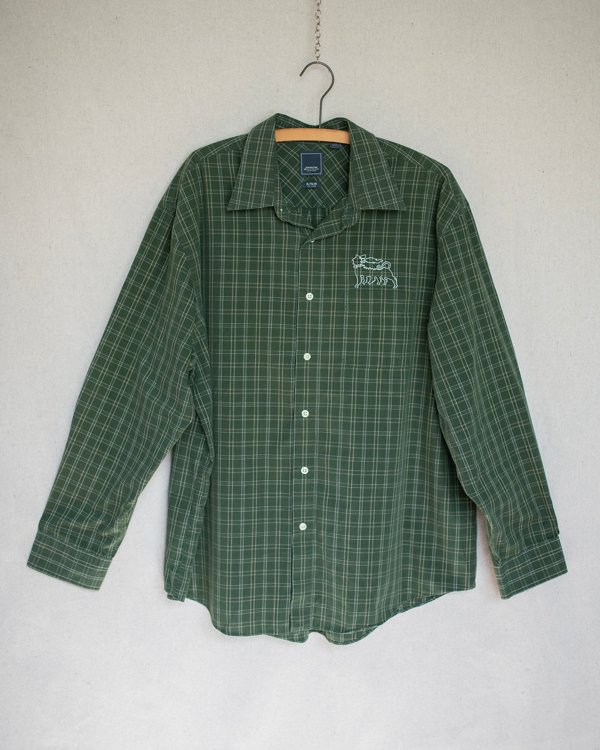 A green long-sleeve plaid button-up shirt hanging on a wooden hanger against a light gray wall. The shirt features a small white embroidered on the left chest.