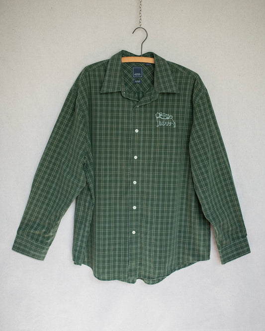 A green long-sleeve plaid button-up shirt hanging on a wooden hanger against a light gray wall. The shirt features a small white embroidered on the left chest.