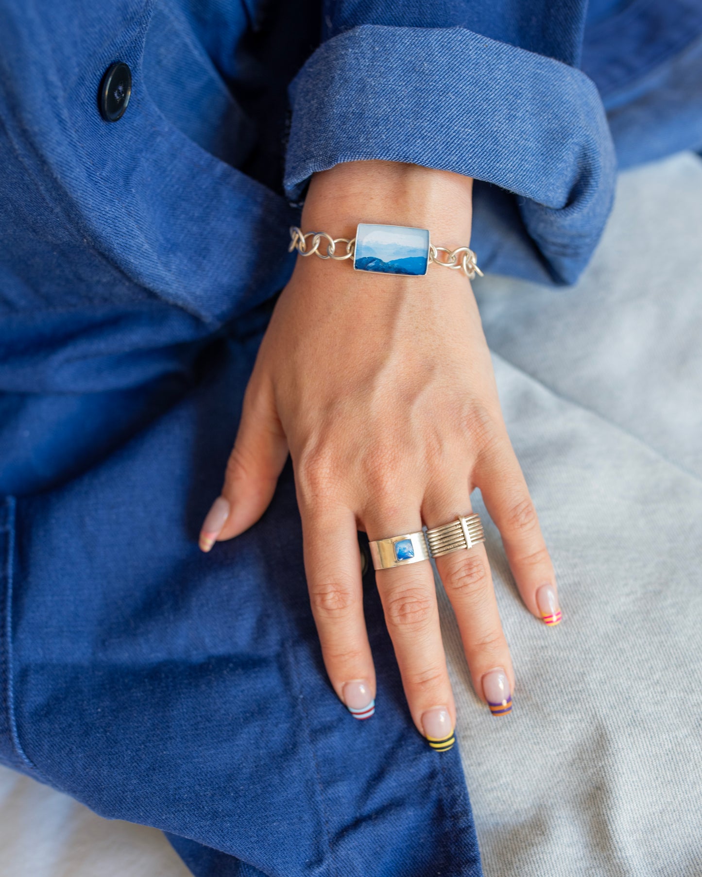 A close-up of a person's hand wearing a silver chain bracelet with a rectangular blue photo charm, along with two silver rings, set against a blue canvas jacket.