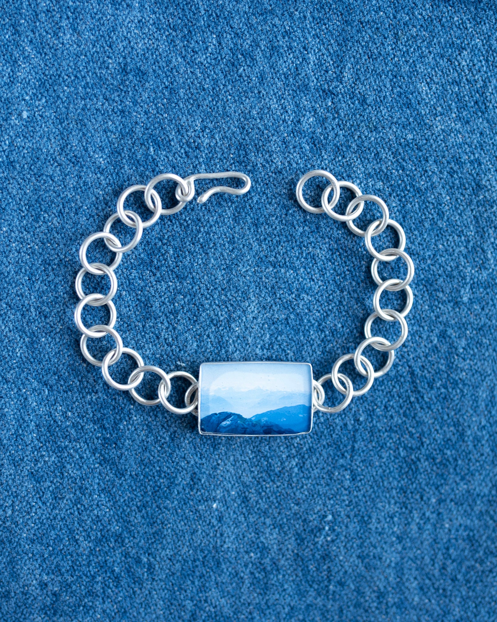 A silver chain bracelet with a rectangular bezel containing a blue and white landscape photo, displayed on a textured blue fabric background.