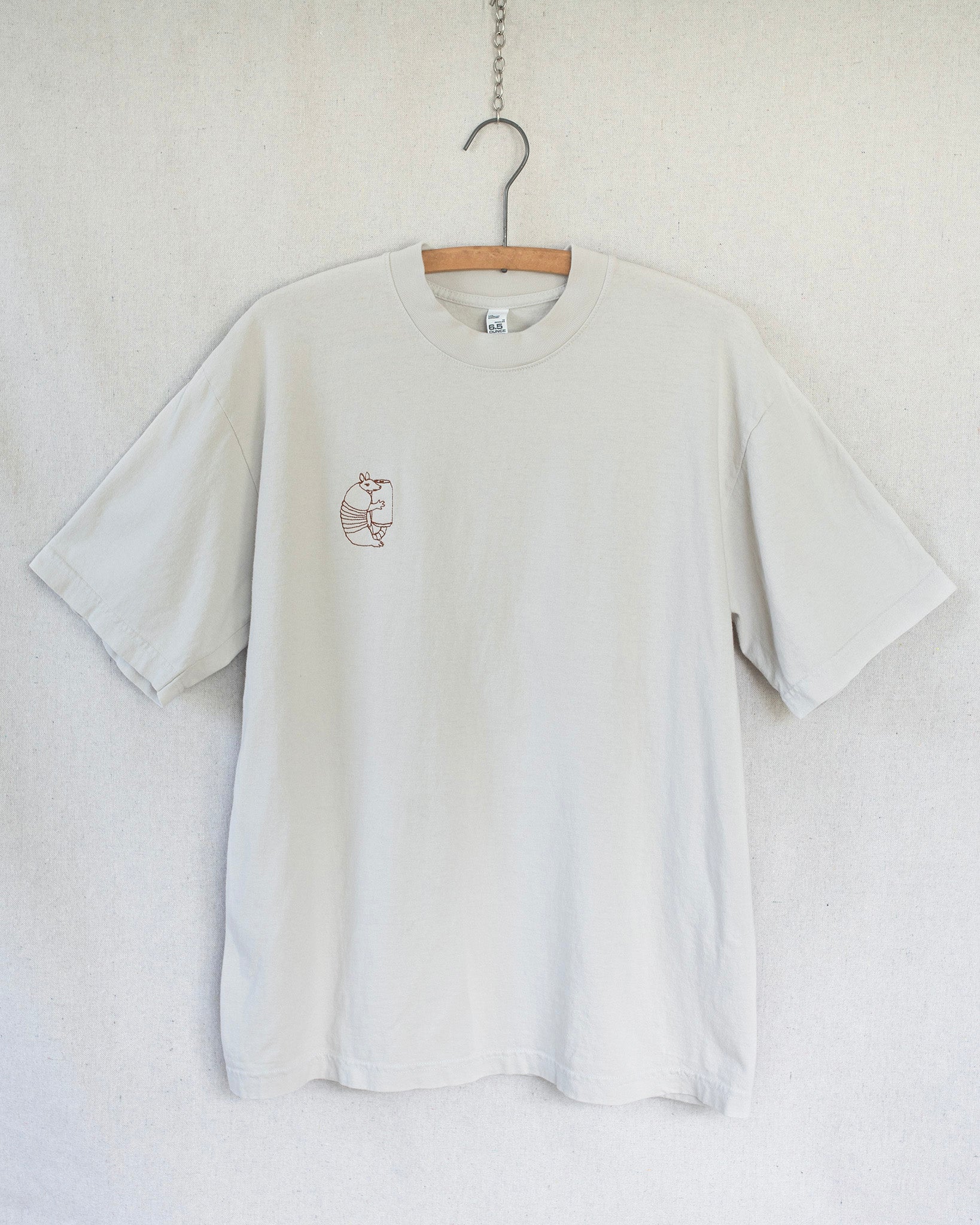A light grey off white crewneck t-shirt hanging on a wooden hanger against a textured off-white wall. The shirt features a small brown graphic of an armadillo on the left chest.