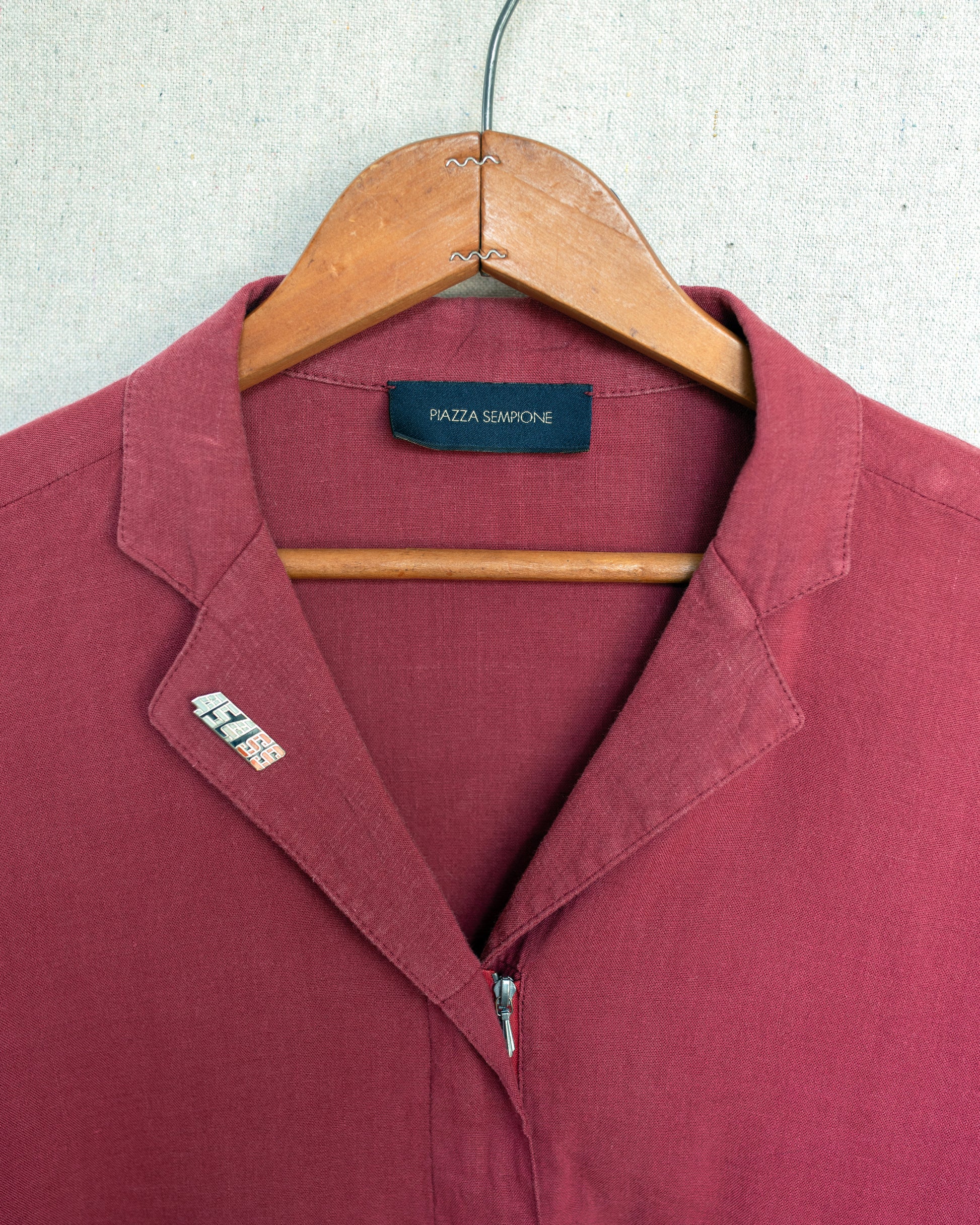 A close-up of a dusty red zip-up shirt on a wooden hanger against a light gray textured background. The brand label inside the collar reads 'Piazza Sempione'.