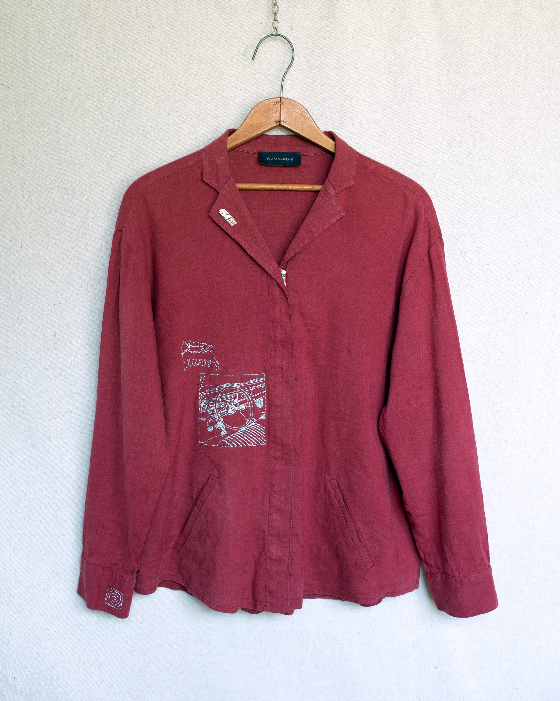 A dusty red jacket with graphic embroideries on the chest, hanging on a wooden hanger against a textured off-white wall.