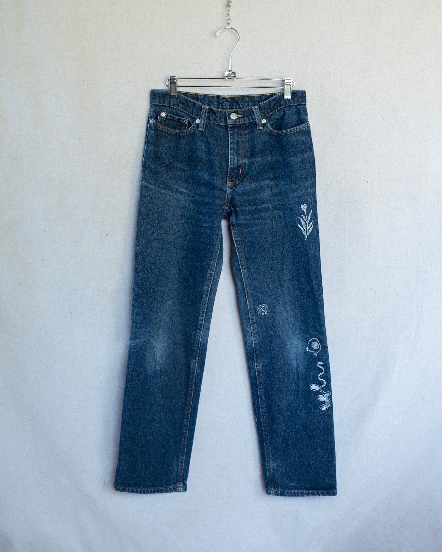 Dark wash low rise jeans with multiple embroideries hanging on a neutral background.