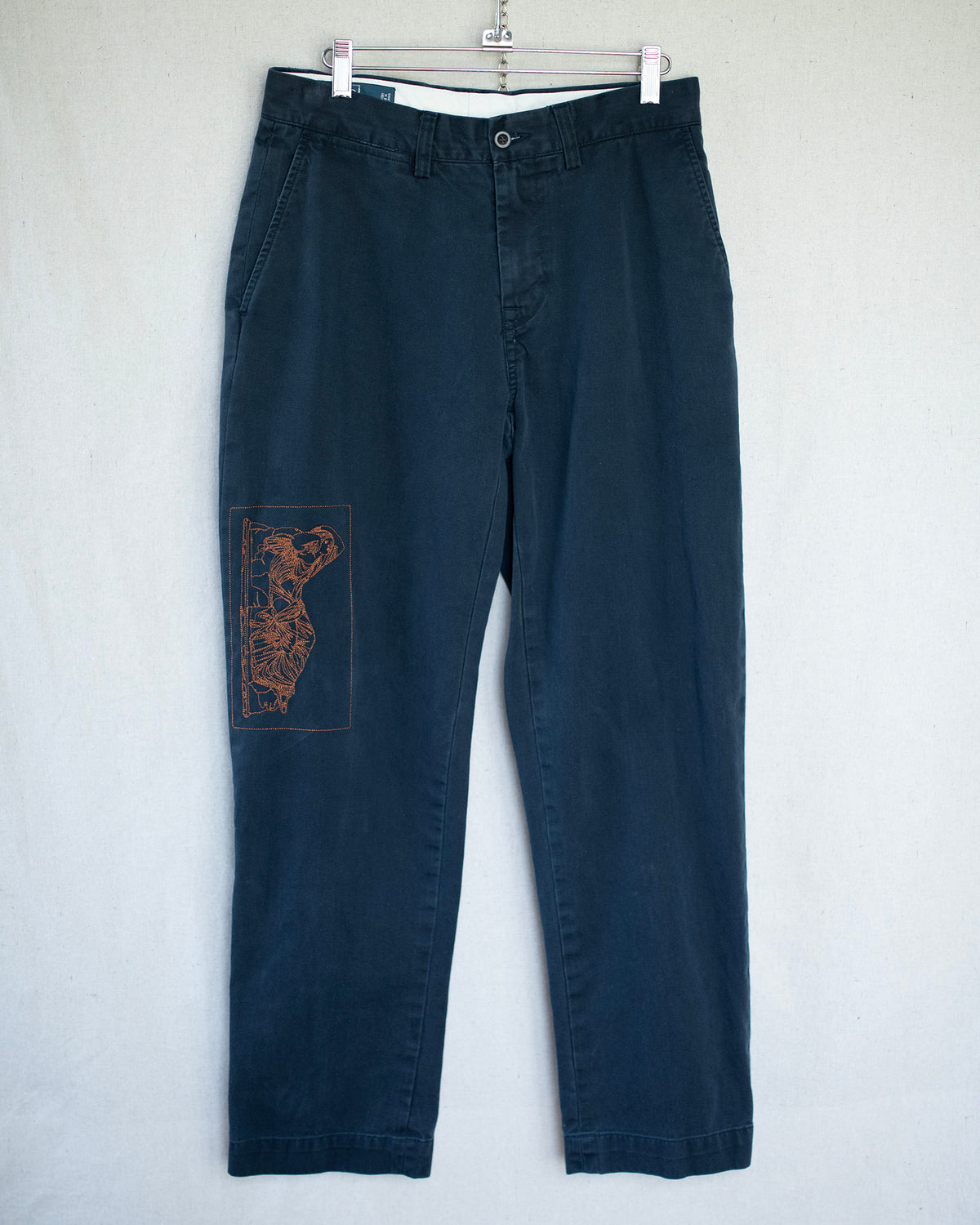 Dark navy blue pants hanging on a white hanger against a light gray wall, featuring a brown embroidered graphic  on the left leg.