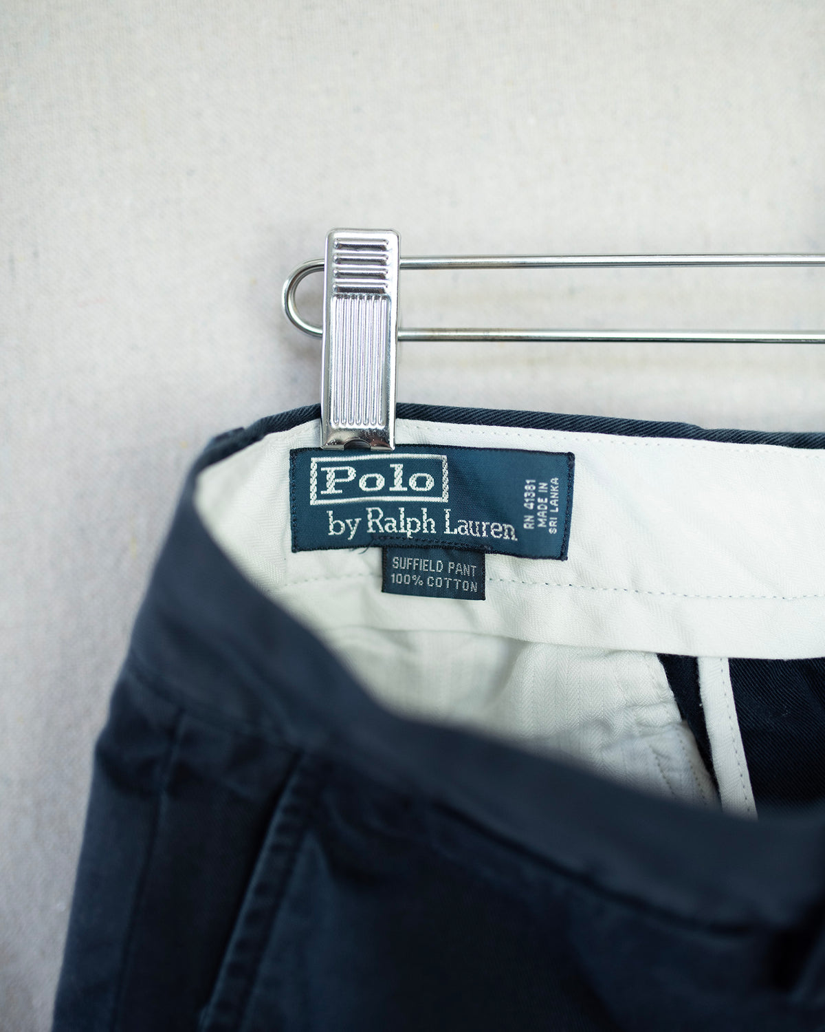 Close-up of Polo by Ralph Lauren navy blue pants hanging on a silver metal hanger against a light gray textured wall.
