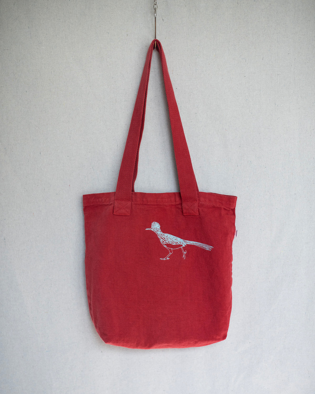 A red canvas tote bag with long shoulder straps, featuring a white embroidered bird design on the front, hanging against a light gray textured wall.