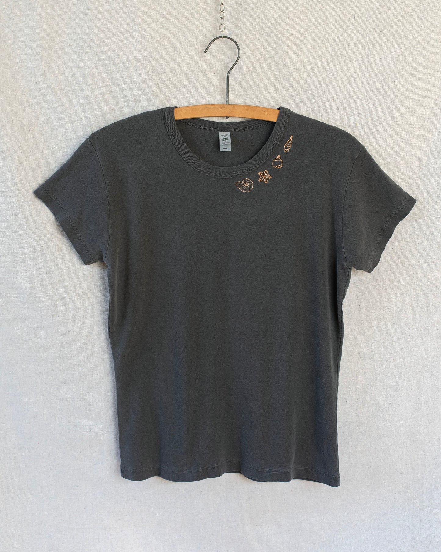 A dark charcoal gray short-sleeve t-shirt hanging on a wooden hanger against a light gray textured wall. The shirt features a small embroidered seashells on one side below the collar.