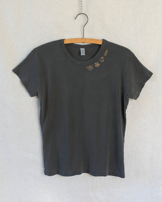 A dark charcoal gray short-sleeve t-shirt hanging on a wooden hanger against a light gray textured wall. The shirt features a small embroidered seashells on one side below the collar.