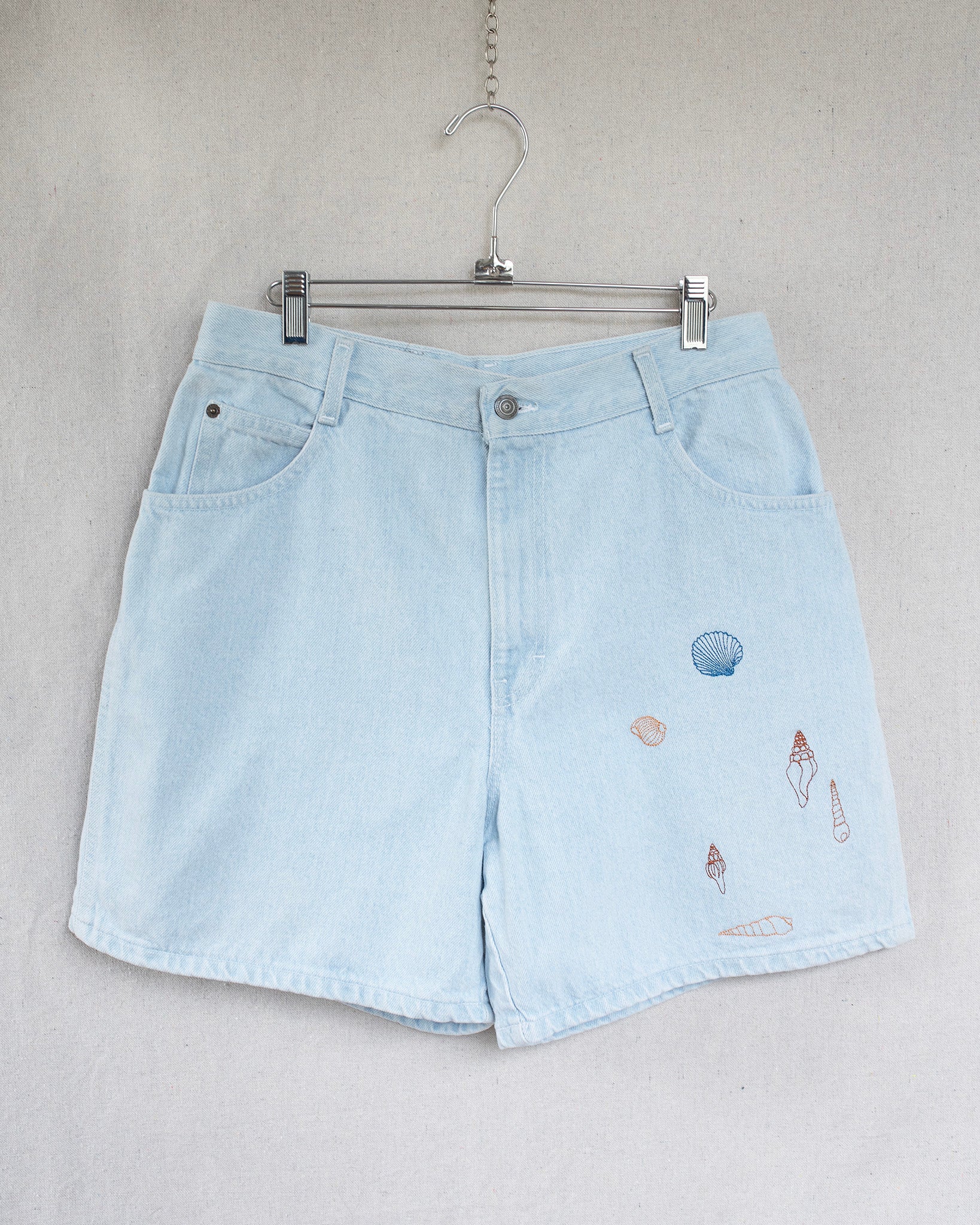 Light blue denim shorts with embroidered seashells on a hanger against a neutral background