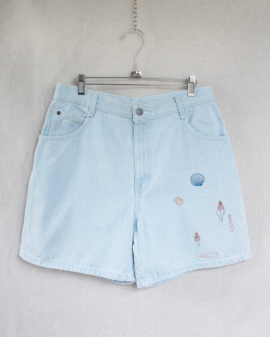 Light blue denim shorts with embroidered seashells on a hanger against a neutral background