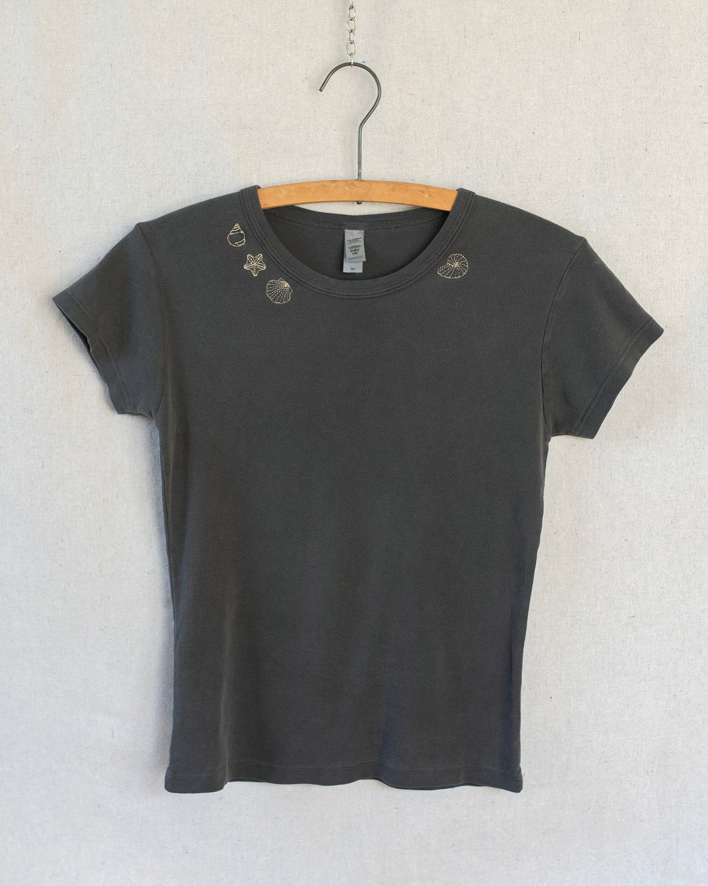 A dark charcoal gray short-sleeve t-shirt with embroidered seashells around the collar, hanging on a wooden hanger against a light textured wall.