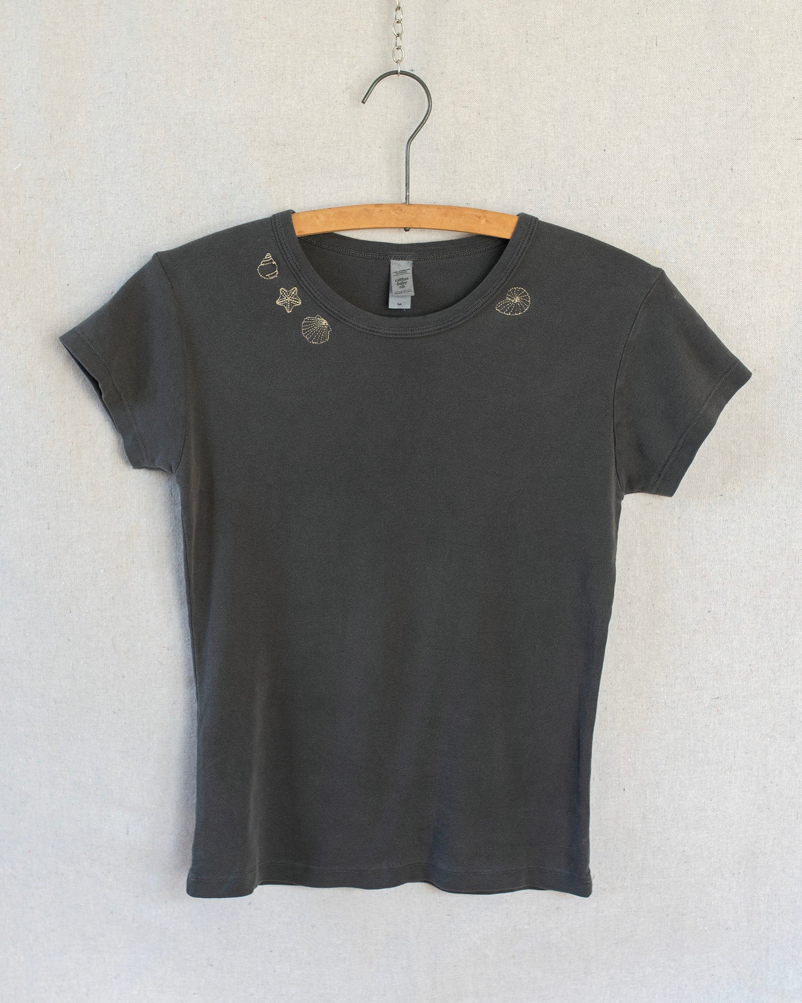 A dark charcoal gray short-sleeve t-shirt with embroidered seashells around the collar, hanging on a wooden hanger against a light textured wall.