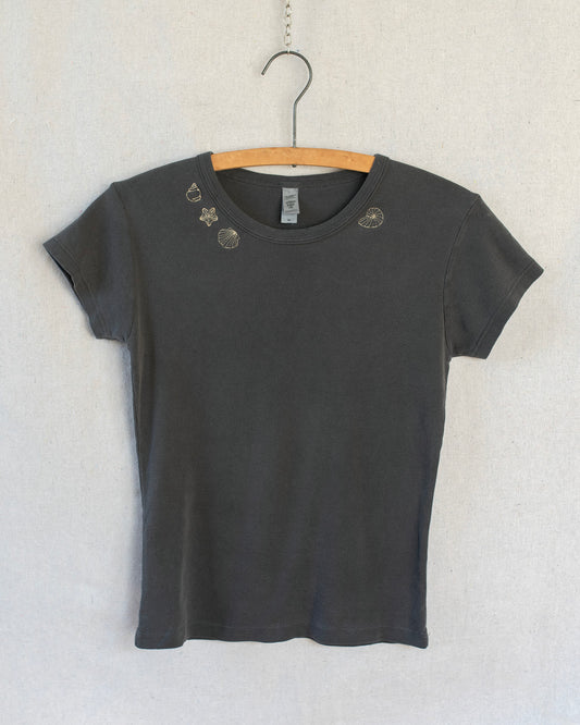A dark charcoal gray short-sleeve t-shirt with embroidered seashells around the collar, hanging on a wooden hanger against a light textured wall.