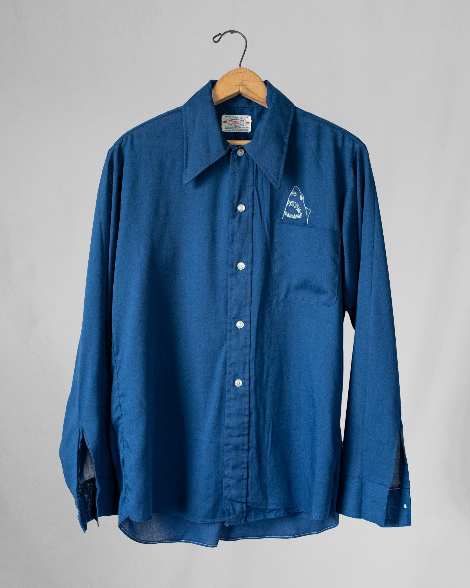 Blue shirt with a shark embroidery over pocket on a hanger against a gray background