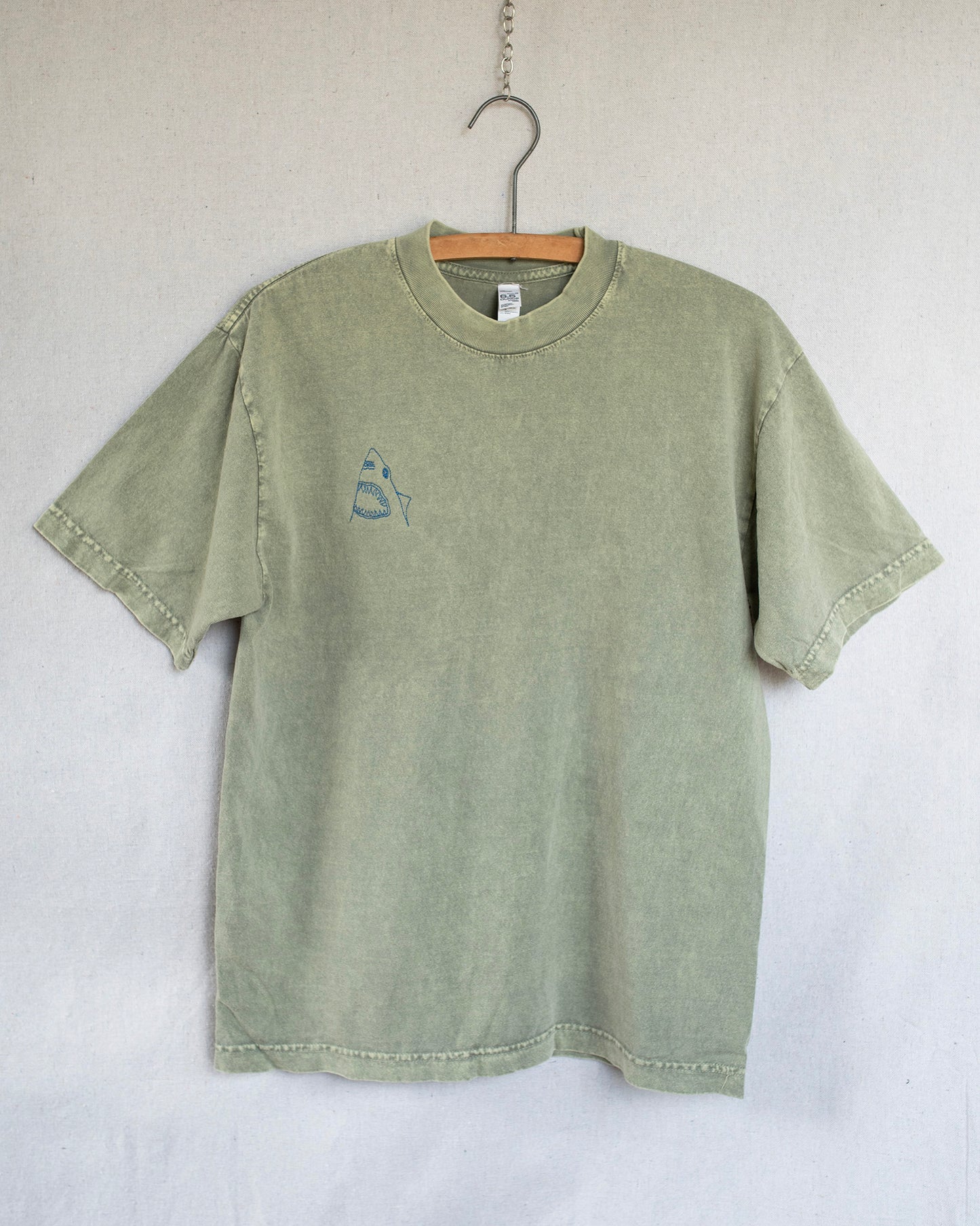 A sage green short-sleeve t-shirt with a small embroidered shark embroidery on the left chest, hanging on a wooden hanger against a light gray fabric backdrop.