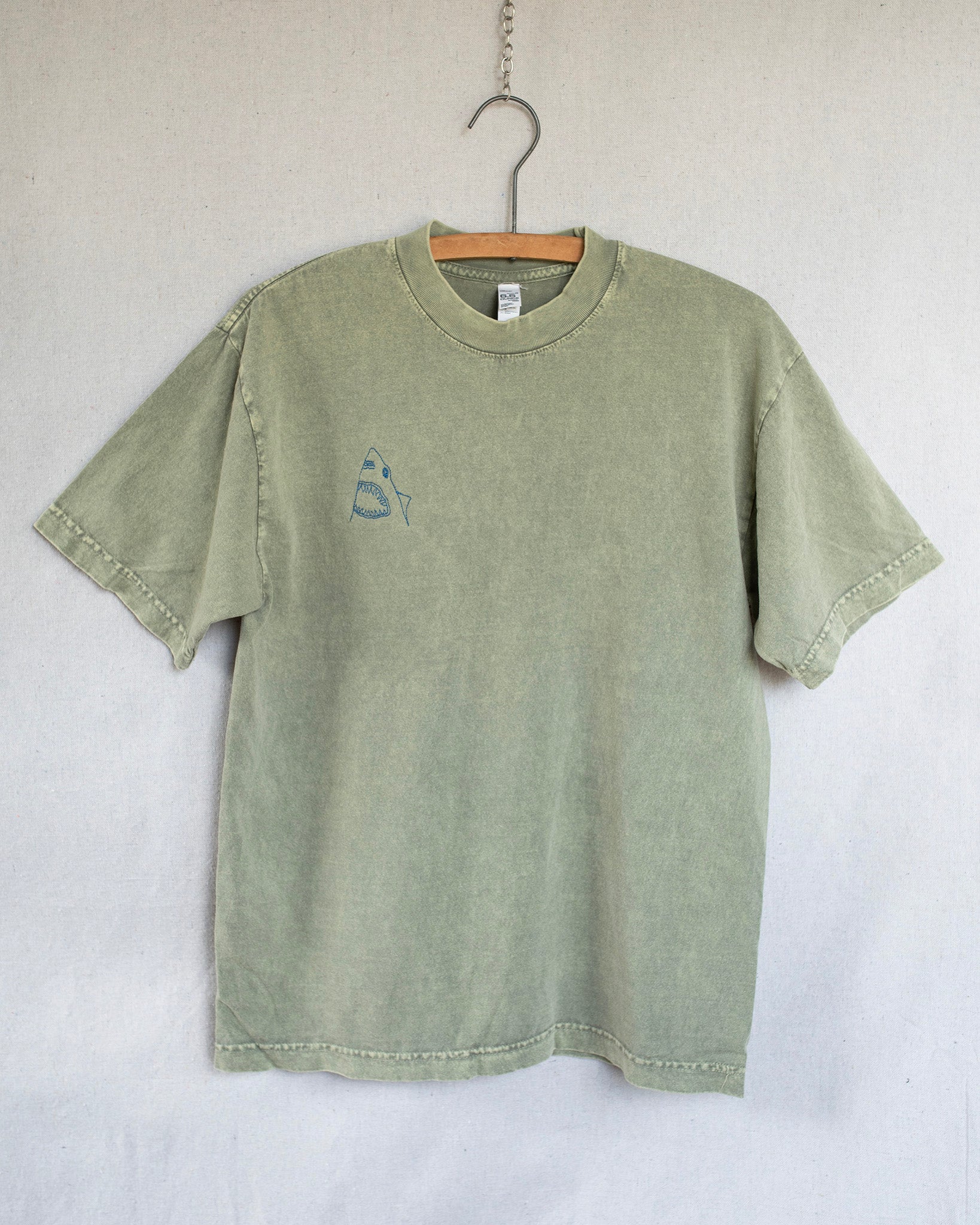 A sage green short-sleeve t-shirt with a small embroidered shark embroidery on the left chest, hanging on a wooden hanger against a light gray fabric backdrop.