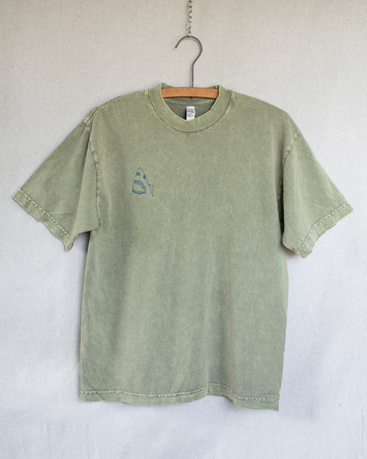 A sage green short-sleeve t-shirt with a small embroidered shark embroidery on the left chest, hanging on a wooden hanger against a light gray fabric backdrop.