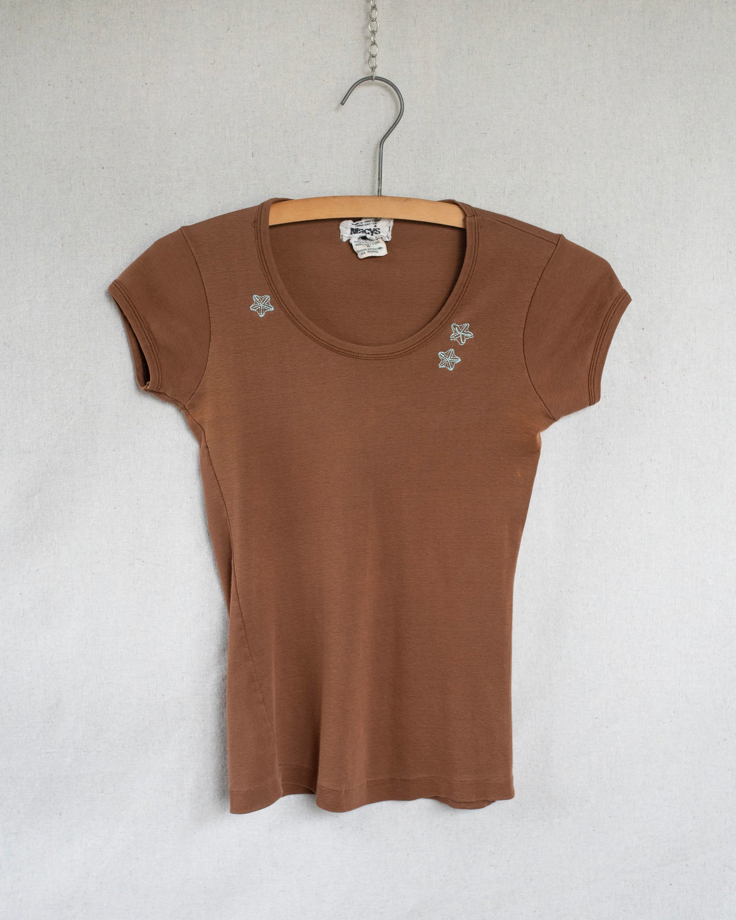 A brown short-sleeve scoop-neck t-shirt with small blue floral embroideries on the chest, hanging on a wooden hanger against a light gray textured wall.