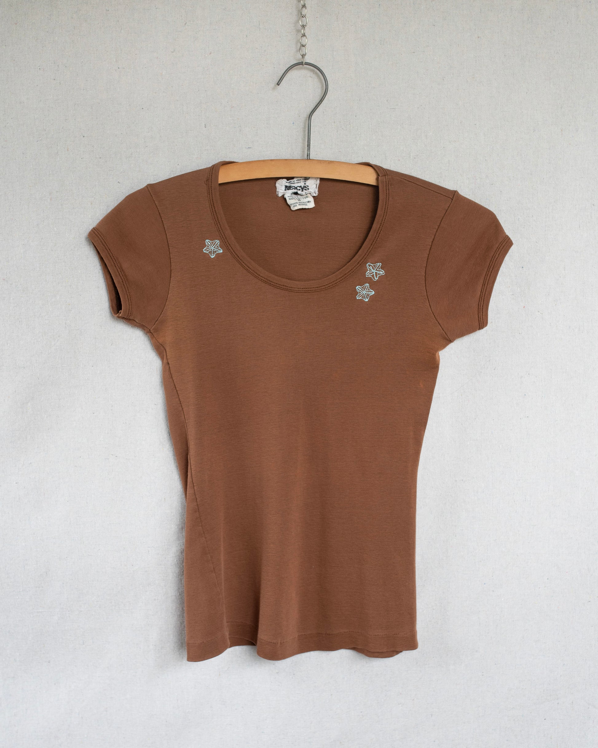 A brown short-sleeve scoop-neck t-shirt with small blue floral embroideries on the chest, hanging on a wooden hanger against a light gray textured wall.