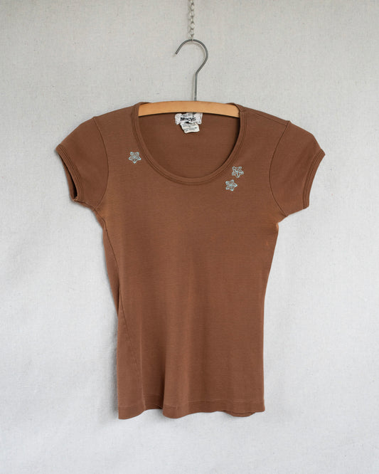 A brown short-sleeve scoop-neck t-shirt with small blue floral embroideries on the chest, hanging on a wooden hanger against a light gray textured wall.