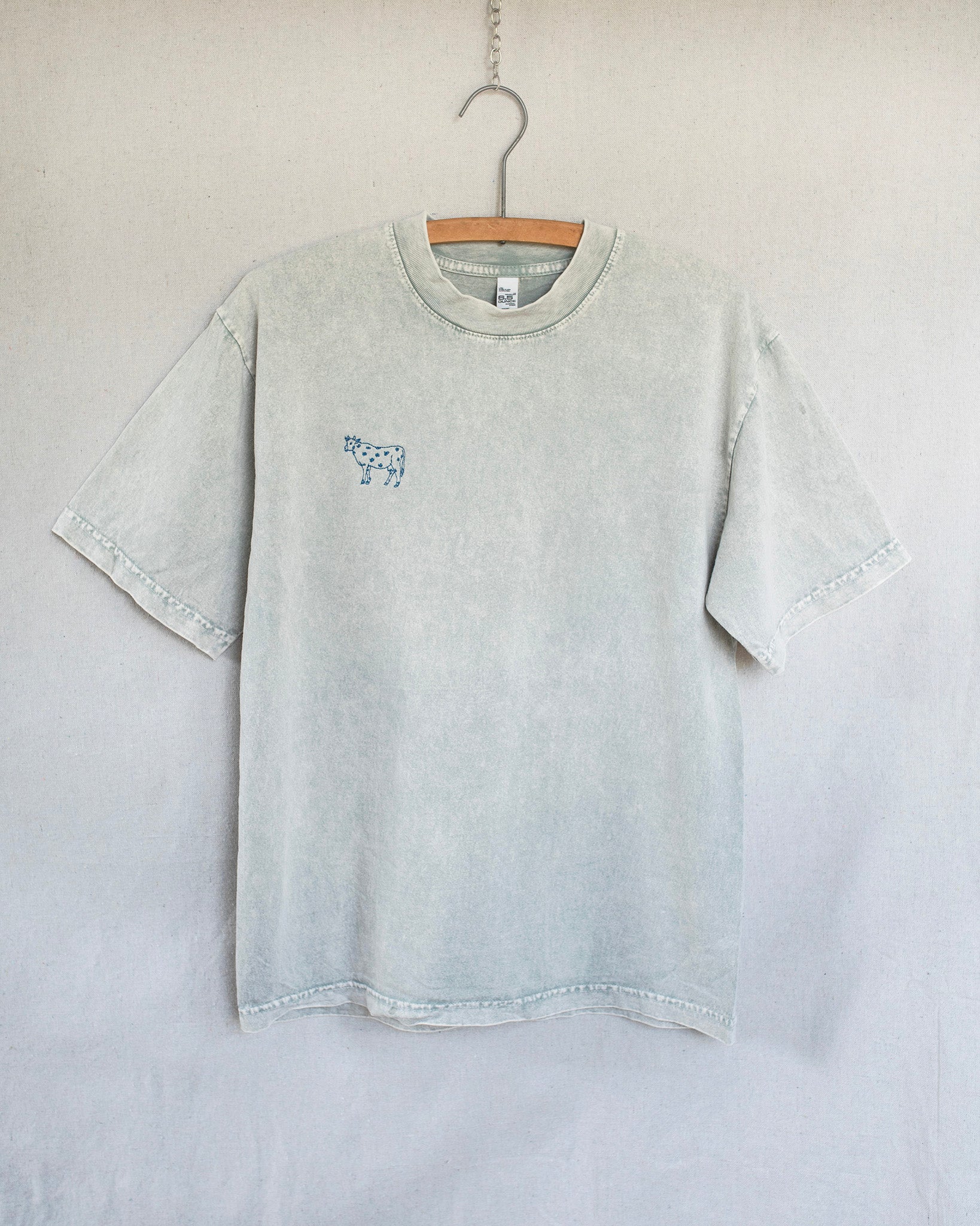 A light gray mineral-wash short-sleeve t-shirt hanging on a wooden hanger against a neutral light gray wall. The shirt features a small blue graphic print on the left chest depicting a cow..