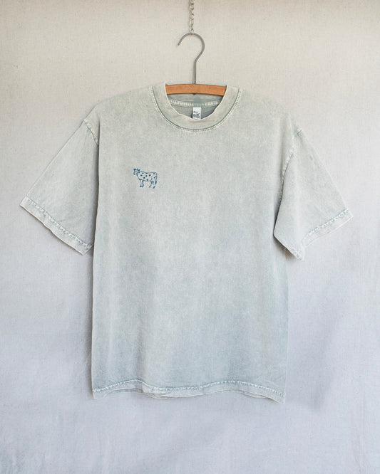 A light gray mineral-wash short-sleeve t-shirt hanging on a wooden hanger against a neutral light gray wall. The shirt features a small blue graphic print on the left chest depicting a cow..