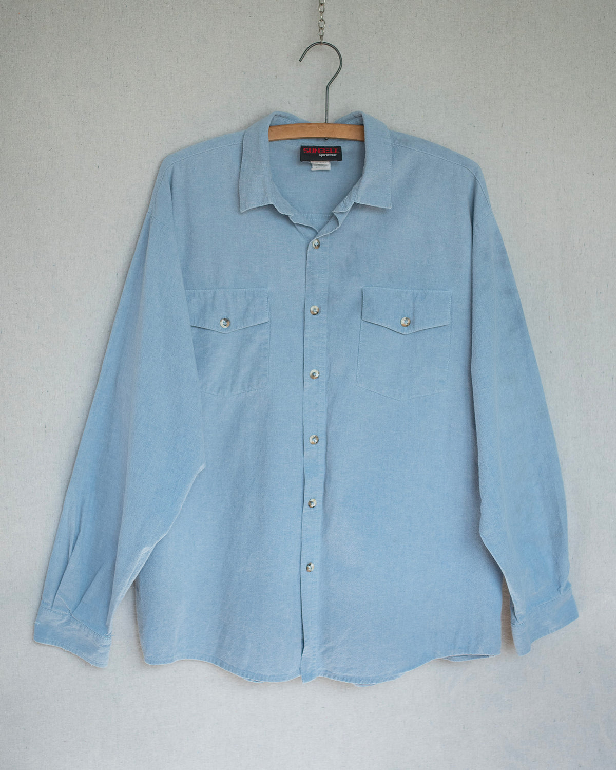 A light blue denim long-sleeve button-up shirt with two chest pockets, hanging on a wooden hanger against a textured light gray wall.