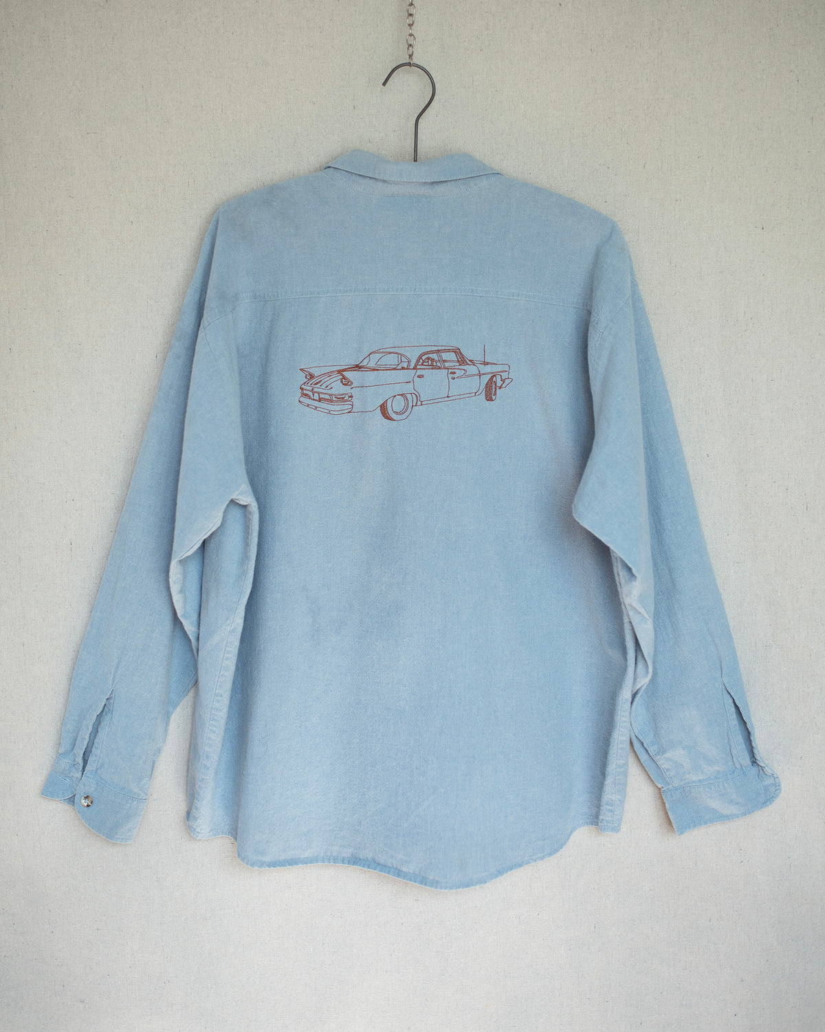 Back of a denim long sleeve that has a car embroidery hanging against a canvas background.