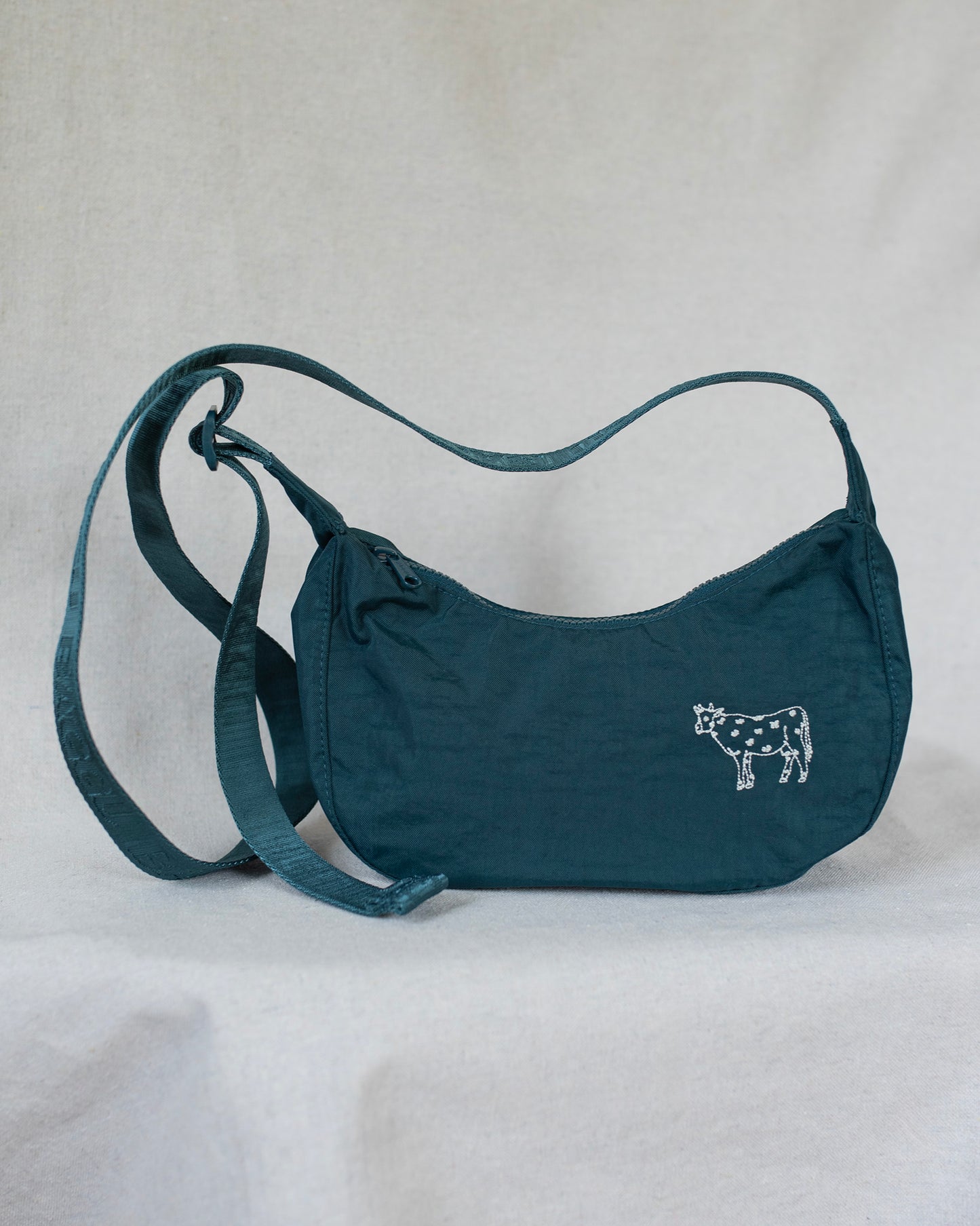 A teal crescent-shaped shoulder bag with a cow embroidery on the front, displayed against a light gray fabric backdrop.