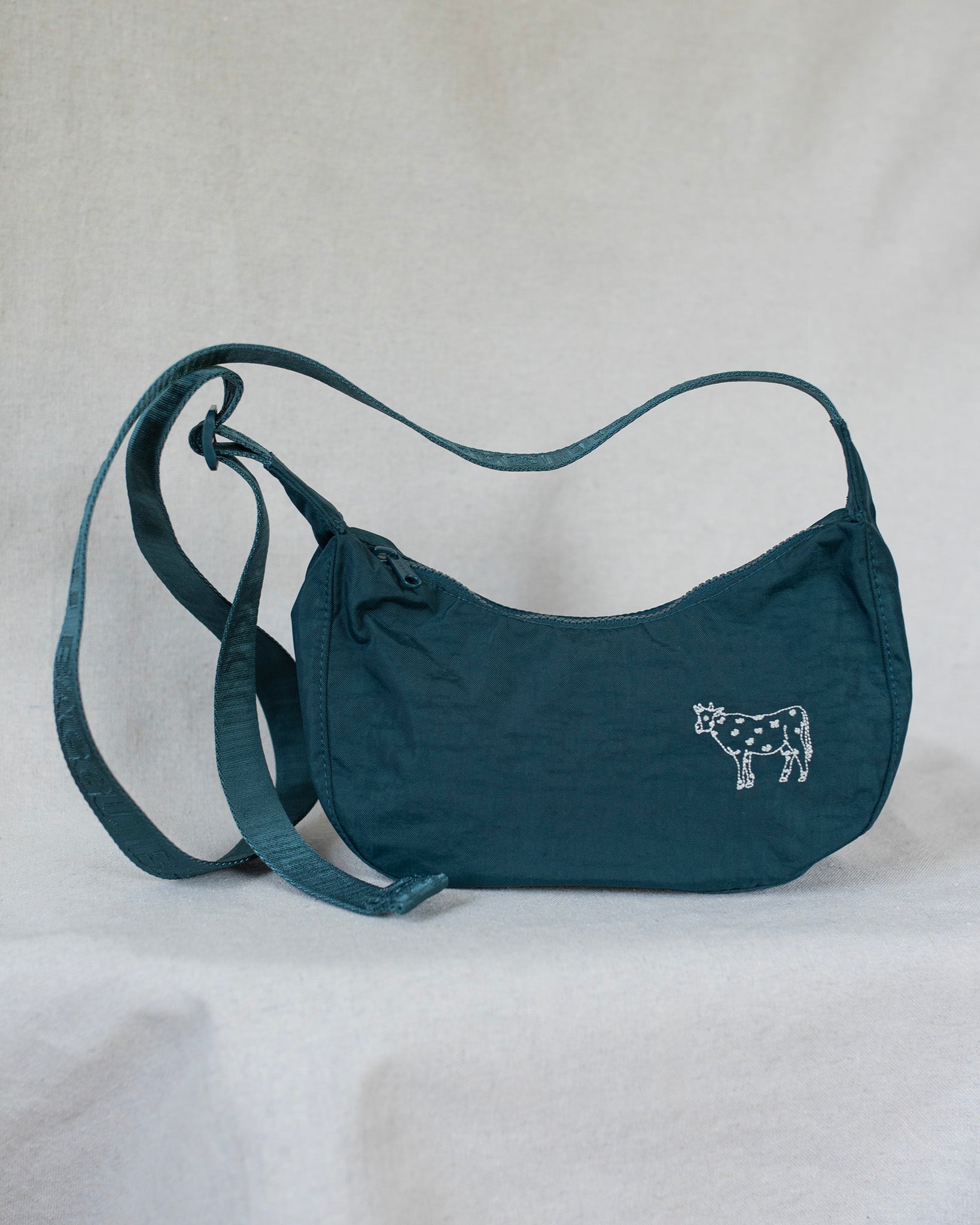 A teal crescent-shaped shoulder bag with a cow embroidery on the front, displayed against a light gray fabric backdrop.