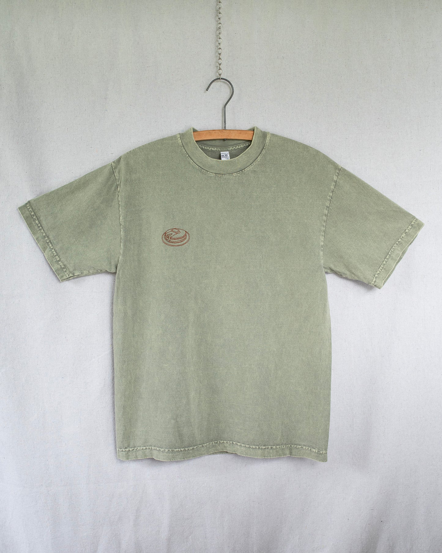 Green t-shirt with a small pancakes embroidery on chest hanging against a white background