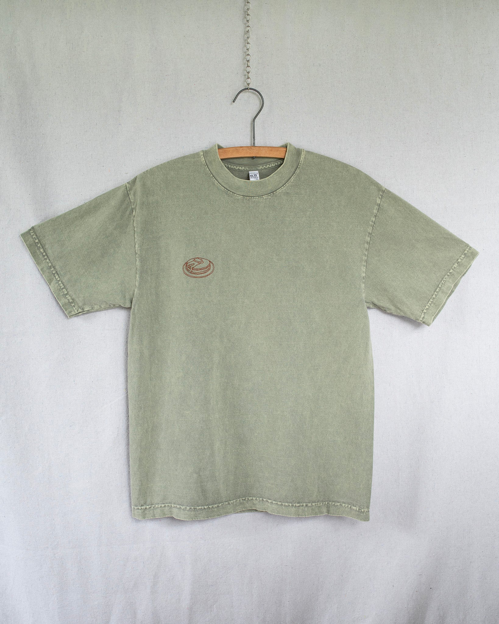 Green t-shirt with a small pancakes embroidery on chest hanging against a white background