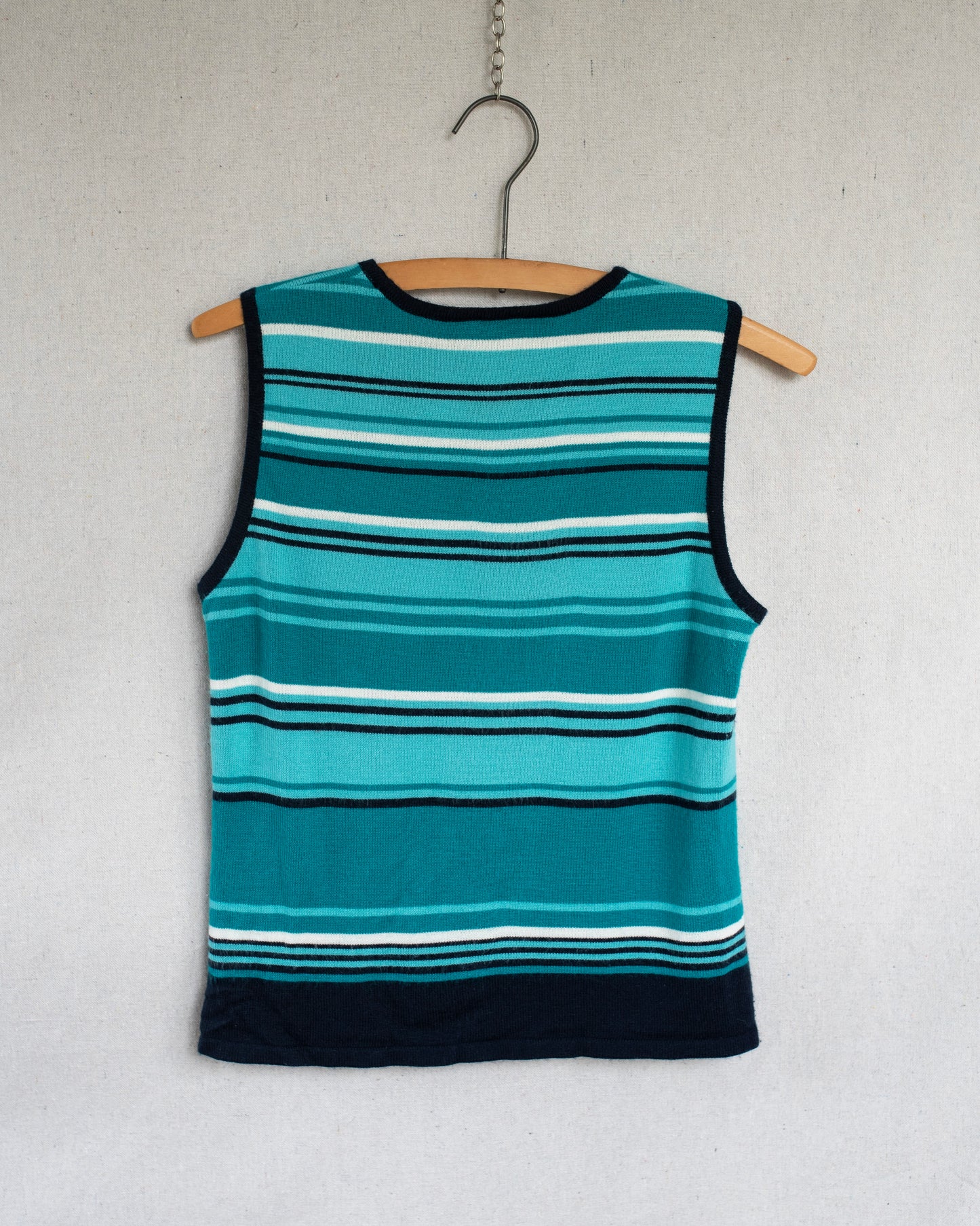 A sleeveless knit top with horizontal teal, blue, white, and black stripes, hanging on a wooden hanger against a light gray textured wall.