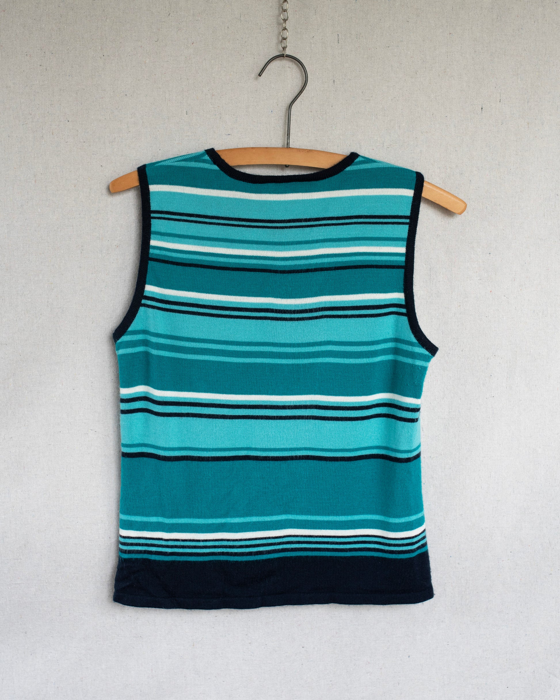 A sleeveless knit top with horizontal teal, blue, white, and black stripes, hanging on a wooden hanger against a light gray textured wall.